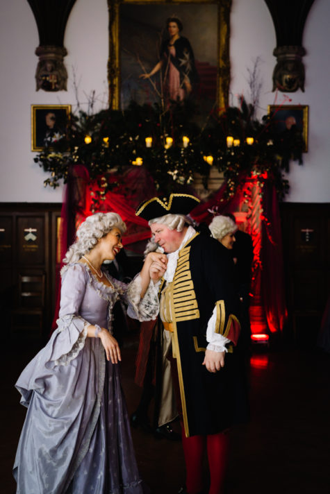 ALT="man and woman in 18th century costumes speak in Chapter Hall"