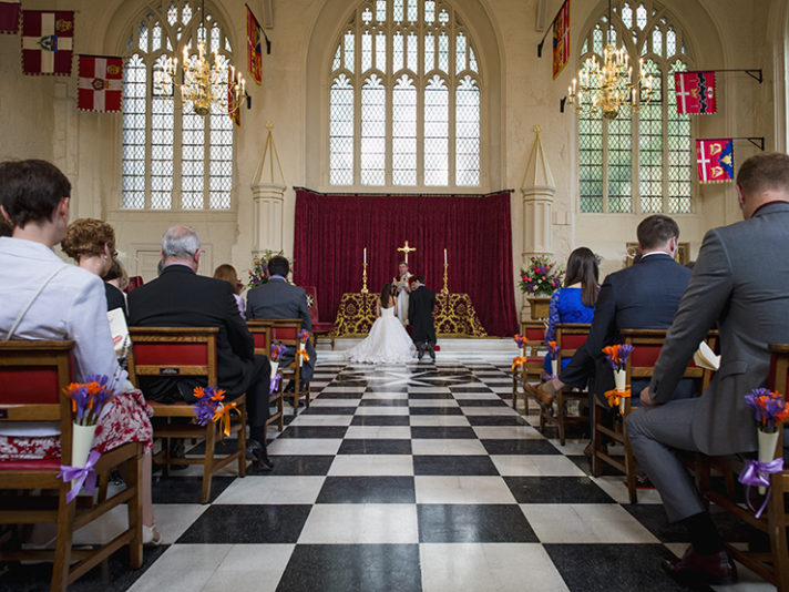 ALT="The aisle of the church is lined with brightly coloured flowers, bride and groom kneel at the altar"