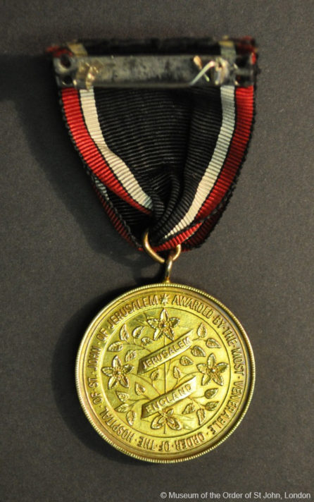 A round gold medal with a central motif of St John's Wort, and writing in a border, suspended from a black, red and white ribbon.