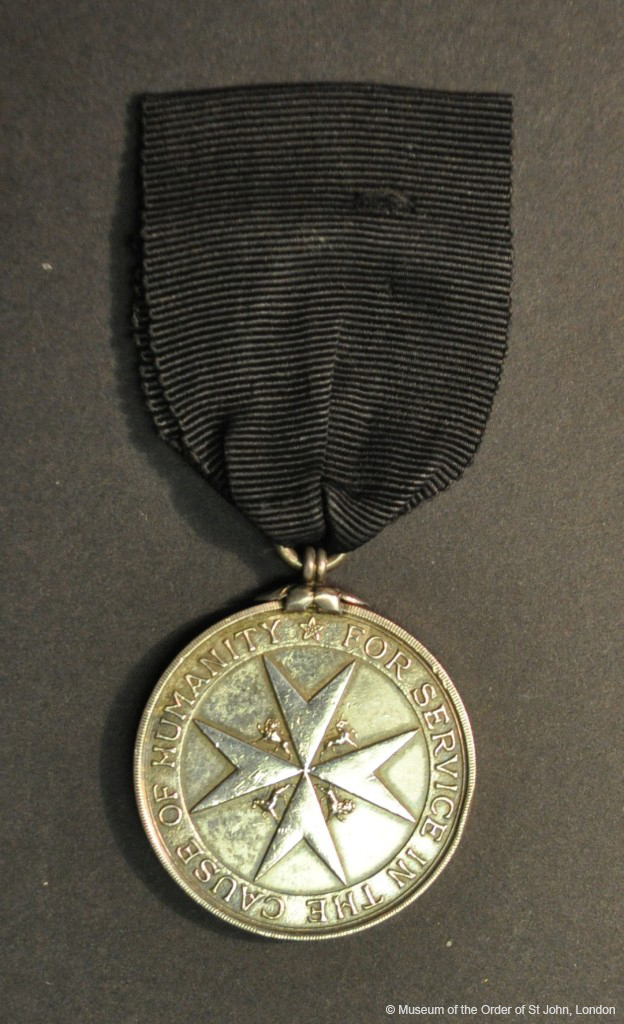 A round silver medal with a central eight-pointed cross with unicorn and lion supporters, and writing in a border, suspended from a black ribbon.