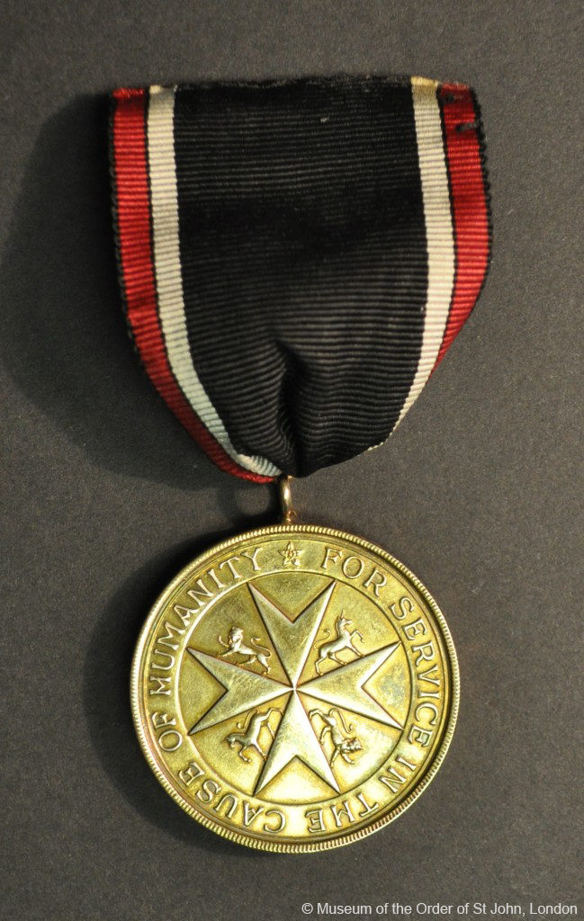 A round gold medal with a central eight-pointed cross with unicorn and lion supporters and writing in a border, suspended from a black, red and white ribbon.