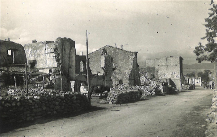 Black and white photograph of half destroyed buildings, many missing walls and roofs, next to a small dirt track road.