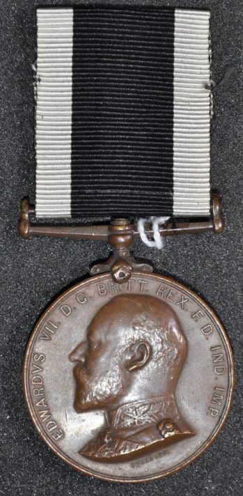 Round bronze coloured medal with a portrait bust. Ribbon has a large black stripe with two thinner white stripes either side.