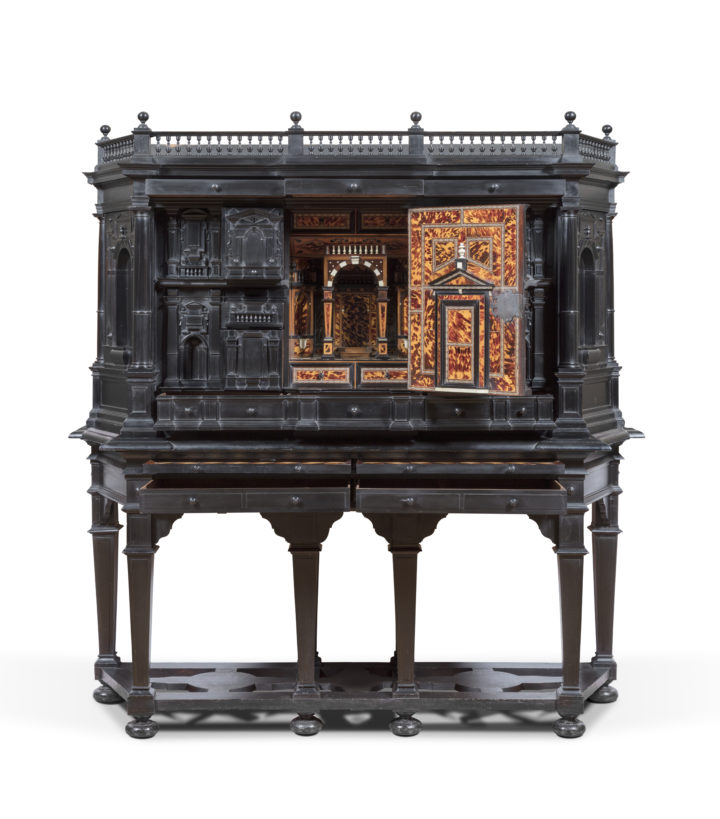 Photograph of a cabinet made of dark wood, standing on a pedestal. A door is open giving a glimpse of an elaborately decorated interior.