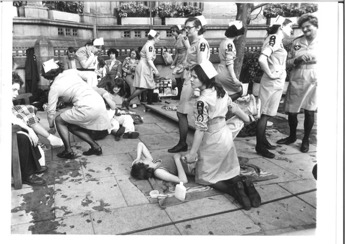 Black and white photograph of St John Ambulance Nursing Cadets in uniform (grey, knee length, belted dresses) attending casualties on a pavement. In the background are casualties sat on benches and in the foreground is a casualty lying on the floor holding her head with a young, concerned looking Nursing Cadet kneeling over her.