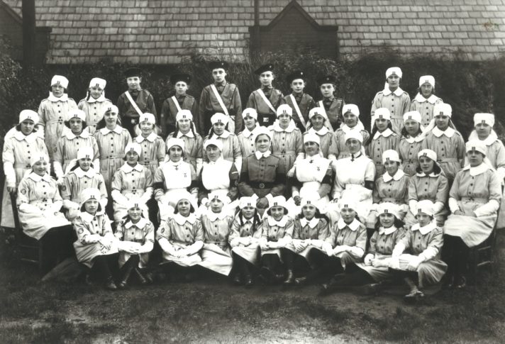 A formal, black and white photograph showing four rows of St John Ambulance volunteers. In the foreground they are sat on the floor and at the back they are standing, possibly on benches. The majority of people photographed are John Ambulance Nursing Cadets wearing grey, button up dresses with white collars and traditional white nursing caps. Seated in the centre is a woman in a matron's uniform, and either side of her sit two women wearing Voluntary Aid Detachment nurse uniforms. At the back of the photograph in the centre are six Boy Cadets wearing black berets, grey shirts and white sashes which run from the right shoulder down to their waist on the left. .