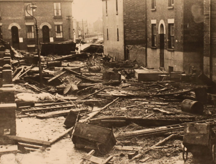 A sepia photograph showing the junction between two streets of terraced houses. Strewn across the road are fences, bins, carts and what appears to be pieces of household furniture including a wardrobe.