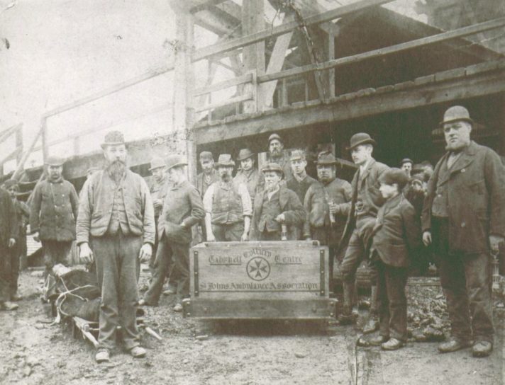 A sepia photograph showing a group of male colliery workers gathered around a wooden cart on which the words 'Tibshelf Colliery Centre St Johns Ambulance Association' are written. All of the men are wearinh waistcoats and bowler hats. To the left of the cart is a casualty on a stretcher presumably from a training exercise. To the right of the cart is a young boy in a flat cap who also appears to be part of the group.