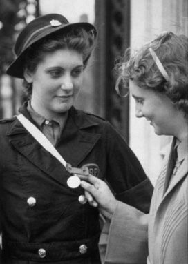 A black and white photograph showing a young, proud looking Betty Quinn in a black ARP uniform jacket and hat. On her chest is a medal which another woman is is looking at smiling.