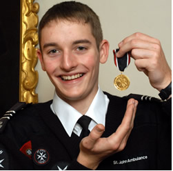 A colour photograph showing a smiling Paul Swift in his Cadet Uniform (a black jumper over a white shirt and black tie) holding up his Gold Life Saving Award in his left hand.