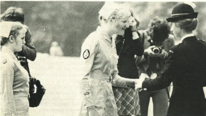 A grainy black and white photograph taken side on of a nursing Cadet wearing a grey belted dress and nursing cap shaking hands with HRH Princess Anne who is wearing black St John Ambulance ceremonial uniform with a black felt hat.