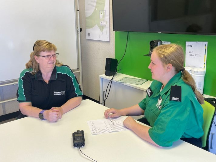 Photograph showing a female Cadet wearing a green service delivery uniform sat at a table with a female St John Ambulance Trainer wearing a black and green polo shirt. On the table between the two women is a recording device