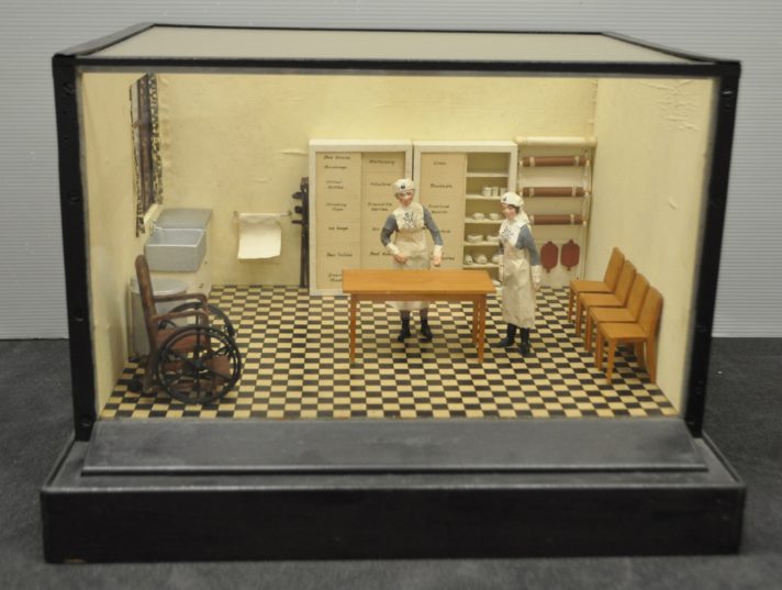 Image of the Nursing Room diorama, glass box with black edges, checkerboard floor with wooden table and two models of nurses wearing St John uniform, shelves behind them with handwritten labels for medical equipment, room also contains a sink, crutches, miniature medical equipment, chairs, a water tank and a small window with floral curtains