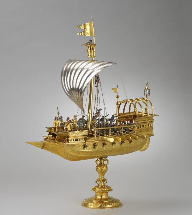 A golden ship model with a silver sail, small figures of soldeirs and oarsmen on the deck.