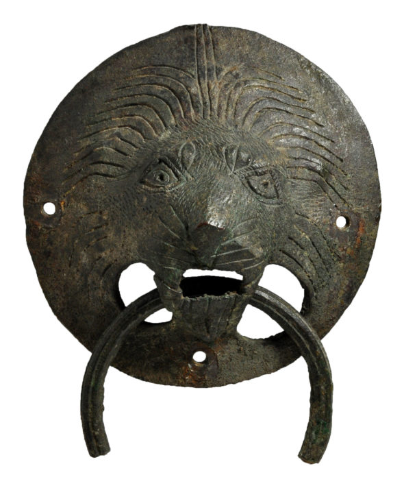 ALT="Bronze lion knocker from stores, circular bronze piece with crudely engraved lion’s face and ring handle attached through mouth"