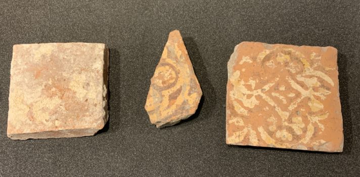 ALT="Three tiles, one on left side is complete with indistinguishable decoration, centre fragment with painted yellow and dark red decorative design, tile on right side is complete with remains of cream and dark red painted design"