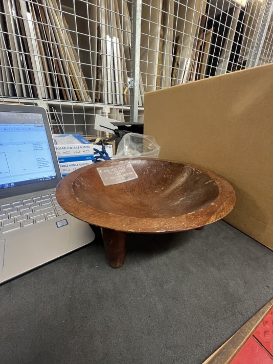 A shallow wooed bowl on legs, photographed on a desk beside a box and laptop.