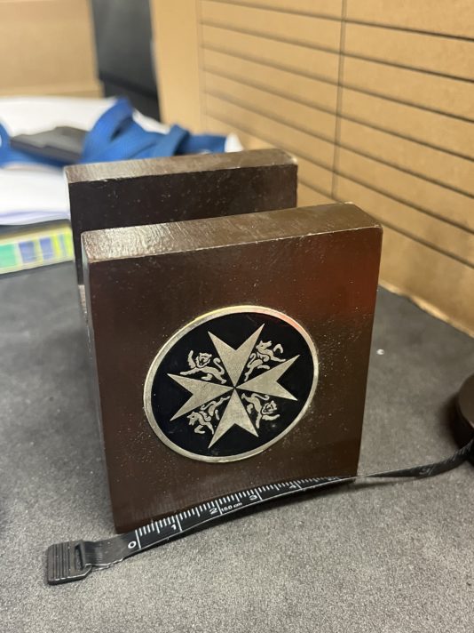 A square wooden item with the emblem of the Order of St John, with a tape measure alongside the object.