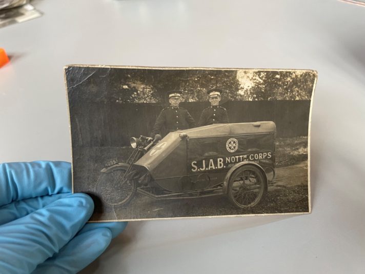 This image shows a gloved hand holding a black and white photograph of two uniformed male Ambulance volunteers standing next to a motorbike sidecar ambulance vehicle.