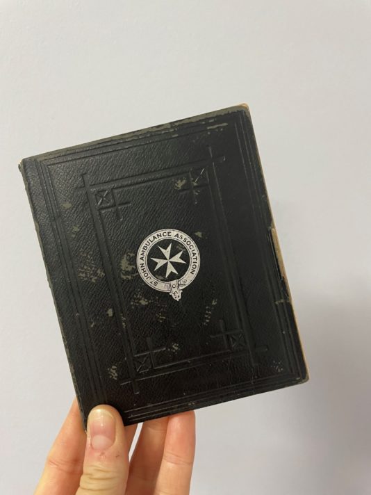 The image shows a small and slim black leather-bound book being held by a hand. In the centre of the cover is the St John Ambulance Association logo of an eight-pointed cross and the words ‘St John Ambulance Association’ embossed in silver.