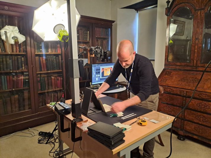 In the library, Dr Steve Gill stands by his desk and carefully handles one of the pamphlets, by placing it on a black mat, leaning on a wedge. Over it, there is a camera fixed to a column, framing the black mat below, whilst behind the desk a photography umbrella ensures the perfect lighting of the room.