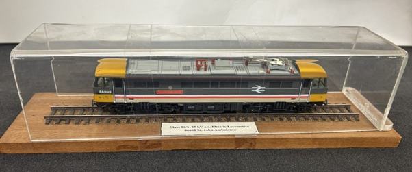 Model of ‘St John Ambulance’ train, on wooden stand with clear plastic case, model inside on railway track. Grey plastic model train with 'St John Ambulance' in red on the side, yellow front and back ends, red details, '86608' in white, white National rail symbol on side.White label in case reading 'Class 86/6 25 kV a.c. Electric Locomotive 86608 St. John Ambulance.