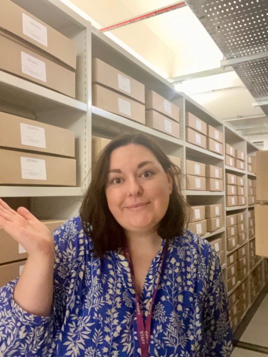 This is a colour photograph of a woman from the waist up, waving and looking towards the camera. She has brown shoulder-length hair and is wearing a flowy blue and white top. She is standing in front of a rack of grey metal shelving which is full of brown cardboard boxes.