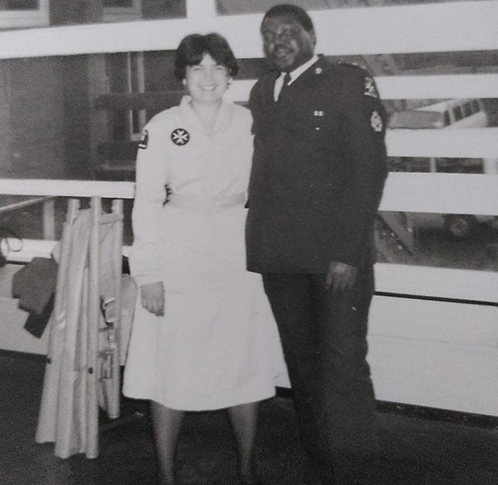 This is a black and white photograph. It shows a smiling young woman in a calf-length light coloured uniform dress standing to the left of a taller man in a dark uniform suit. They are inside a building with windows behind them.