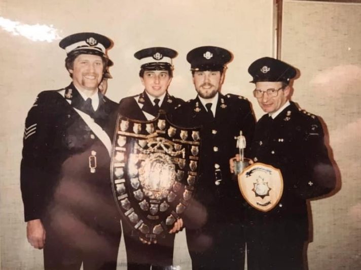 This is a colour photograph of four men looking towards the camera. They are wearing black uniform suits with white shirts, black ties, and black caps. Between them they are holding a large wooden shield with 30 small silver plaques on it, and a smaller wood and gold plated shield.