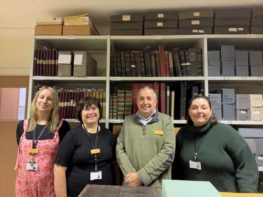 This is a colour photograph of four people (three women and one man) smiling and looking towards the camera. Behind the people is a metal shelving unit filled with books and boxes. The people are dressed in their own clothes, and each wears a name badge.