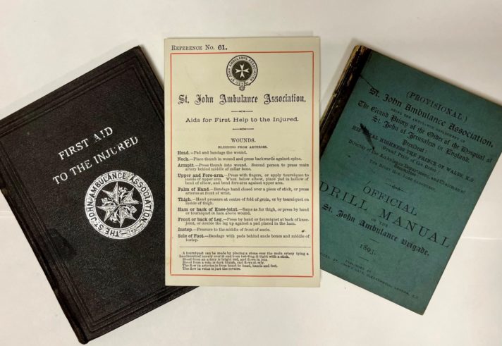 Three fanned St John Ambulance Association printed manuals with titles 'First Aid To The Injured,' Aids for First Help to the Injured (Wounds),' Official Drill Manual'