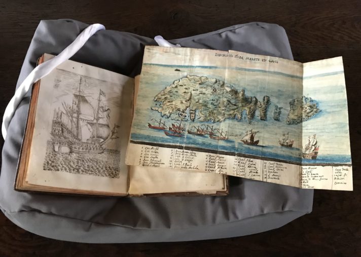 An open book rests on a cushion with some weights holding it open, accompanied by a previously folded sheet. The left-hand page shows a seventeenth-century ship, whilst on the sheet we find an illustrated map of Malta and Gozo.