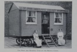 A black and white photograph of two women dressed in light long dresses and light tops and seated in front of a large wooden structure on wheels. The structure is a mobile hospital, with two windows either side of the door.