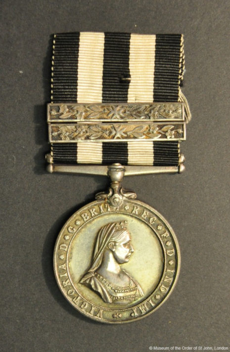 A round silver medal with he side profile of Queen Victoria. It is attached to a ribbon with 5 black and white vertical stripes. Attached to the ribbon are two silver horizontal bars.