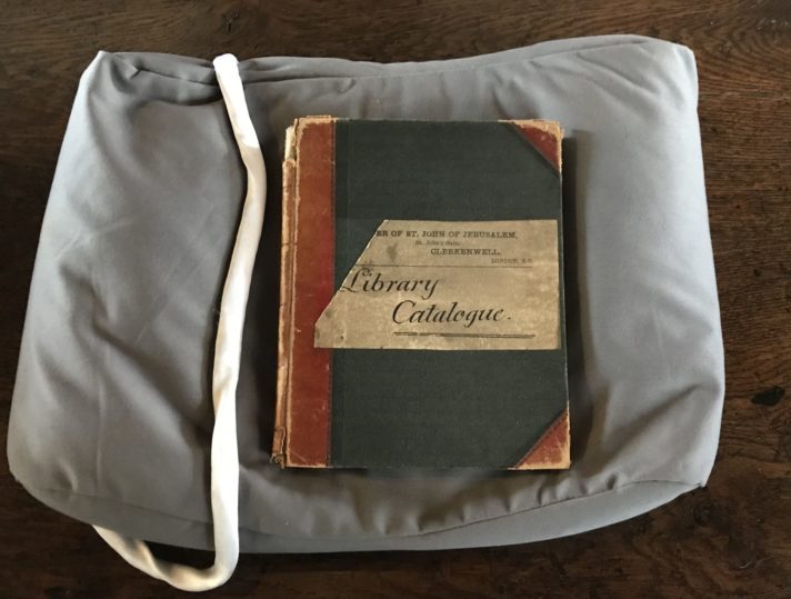 A closed book rest on a cushion with a weight nearby. The label on the binding reads ‘Library Catalogue’.