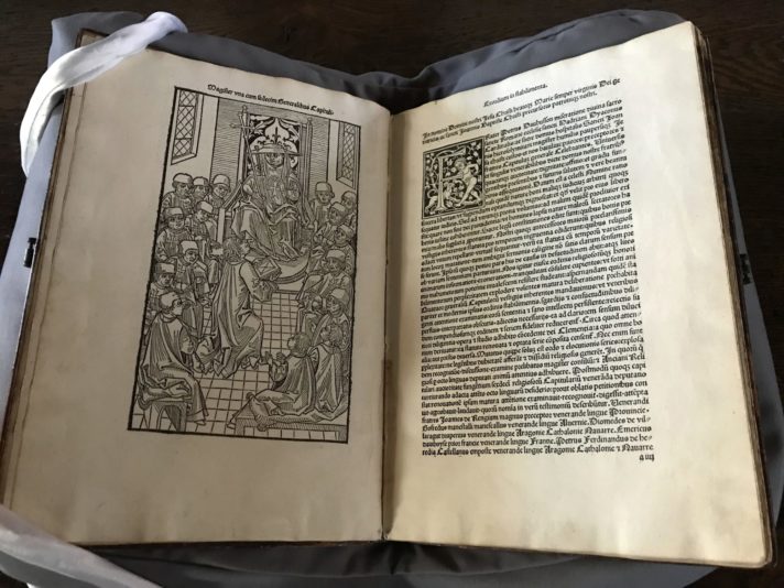 An open book rests on a cushion with some weights holding it open. On the left-hand page, a full-page woodcut shows a group of knights hospitallers, with one of them offering a book to their Grand Master. On the right-hand side page, there is the beginning of the text.
