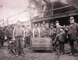 A black and white photograph of a group of men and some small boys standing in front of a mine. Amongst the men is a wooden trolley with the words 'Tibshelf Colliery Centre'.