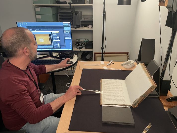 A man sitting at his desk looks at a screen on his side, checking the photograph of a manuscript page he has just taken. In front of him, a manuscript volume is placed on a black mat on his desk. Over it, there is a camera fixed to a column, framing the black mat below.