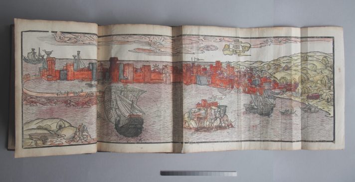 A fold-out page from the book shows a woodcut illustration hand-coloured in yellow, red and brown. It depicts a view of the harbour of Methoni with two docked ships and one sailing in the distance.