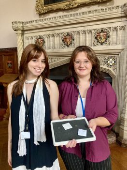 Two white women with brown hair stood in front of the fireplace in Chapter Hall holding out a black tray containing multiple coins.