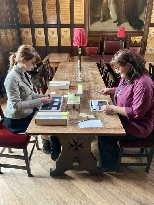 Two white women with brown hair sat at a wooden table in the panelled Council Chamber room. They are working with the coins on the table in front of them. 