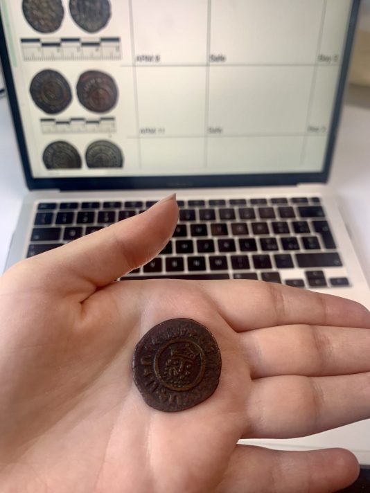 The foreground features Ilenia's hand holding a round crusader coin in her palm. In the background is her laptop open at a spreadsheet of the coin collection audit. 