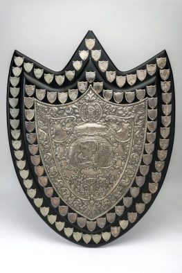 A black painted wooden shield. The central shield plaque has a highly decorative impressed decoration featuring a central image of a horse-drawn ambulance in the background and people in the foreground, with five male uniformed officers tending to a patient on stretcher. Surrounding the central plaque is a border of 40 small shields dating from 1904-1954, with another border of 55 shields dating from 1955-2000.