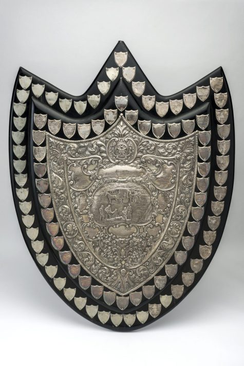 A black painted wooden shield. The central shield plaque has a highly decorative impressed decoration featuring a central image of a horse-drawn ambulance in the background and people in the foreground, with five male uniformed officers tending to a patient on stretcher. Surrounding the central plaque is a border of 40 small shields dating from 1904-1954, with another border of 55 shields dating from 1955-2000.