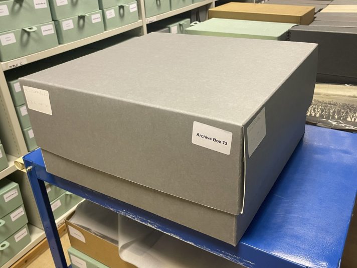 A large grey cardboard box, labelled with a white box number label, sits on top of a blue metal trolley. In the background are smaller green labelled cardboard boxes on grey metal shelving.