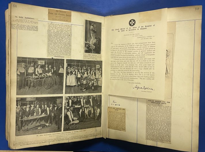 A large scrapbook lies open on a blue table. The pages contain cuttings of textual articles with some accompanying black and white photographs from old newspapers. Also included are pasted in letters.