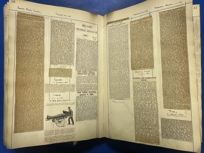 A large scrapbook lies open on a blue table. The pages contain cuttings of textual articles with some accompanying black and white photographs from old newspapers.