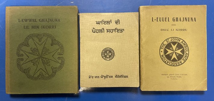 Three small books lie closed, flat, and in a line on a blue metal table. The book on the left is green, and the books in the middle and on the right are beige. They are first aid manuals for St John Ambulance. Each manual has the title in black at the top of the cover, and has the logo of St John in the centre – the eight pointed cross – in black.