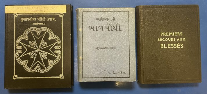 Three small books lie closed, flat, and in a line on a blue metal table. The book in the middle is a light blue, and the books on either side are black. They are first aid manuals for St John Ambulance. Each manual has the title at the top of the cover, either in black or white text, and the manual on the left has the white logo of St John in the centre – the eight pointed cross – in black.