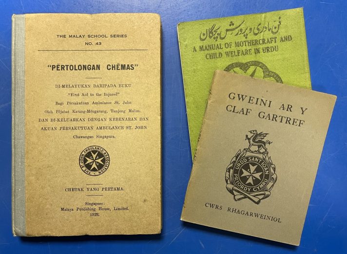 Three small books lie closed and flat on a blue metal table. They are first aid manuals for St John Ambulance. Each manual has the title at the top of the cover, either in black or white text, and the manual on the left has the white logo of St John in the centre – the eight pointed cross – in black.